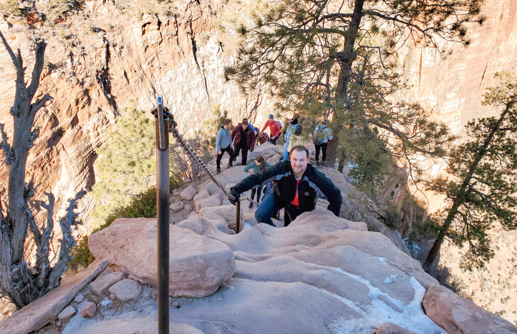 A man holds onto a chain leading up a very skinny path with natural cared rock steps leading up