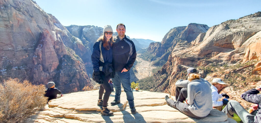 A man and woman stand on the edge of Angel's Landing looking out at the large gorgeous bright orange canyon