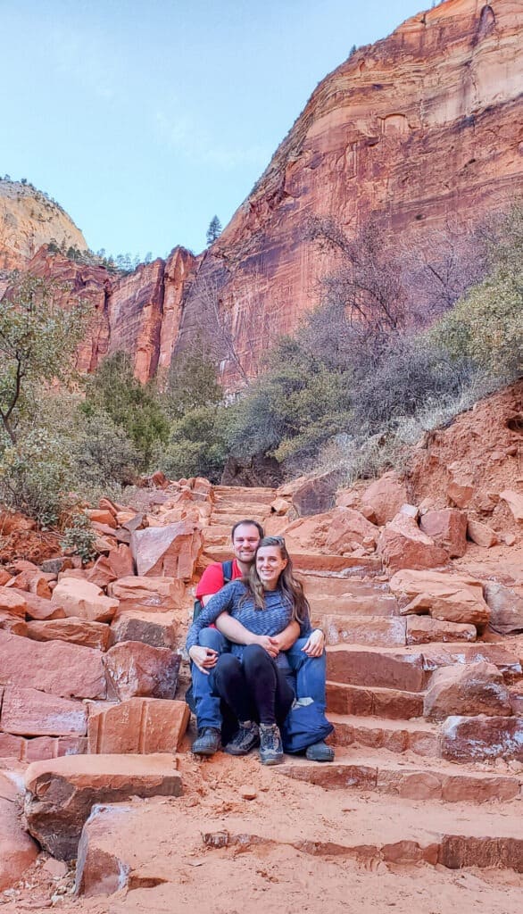 A man and woman sits on the light orange rock steps leading up the Emerald Pool hike