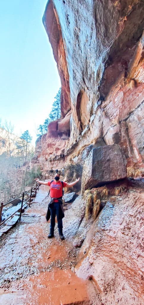 A man in a red jacket stands on the wet and muddy rock path leading around the large tan rock wall with multiple people ahead on the path