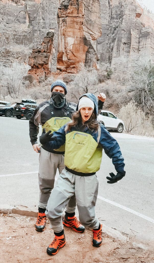 A man and woman in water proof green and black dry-suits in a cement parking lot on a mountain.