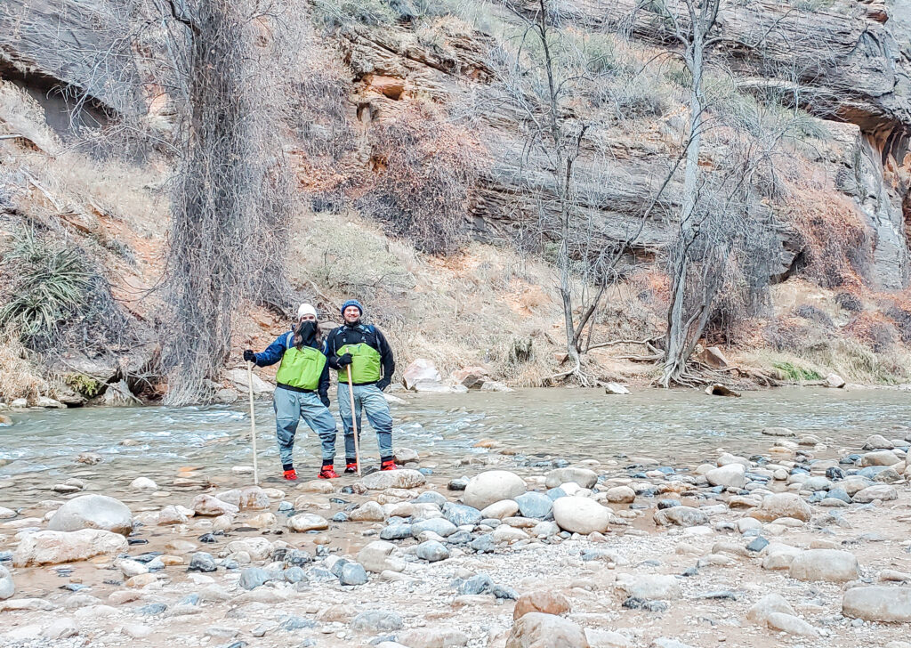 A man and woman on the rocks of the bank of the narrows ( a river). The river is located at the base of a yellow/sandy mountain with dead trees.