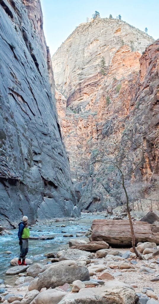 A woman stands in a blue water stream looking up at a large black and orange rock wall