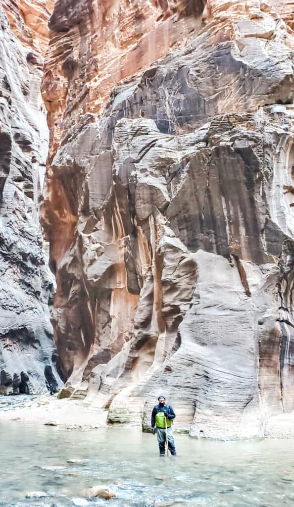 A man standing (in ankle deep water in the middle of a river) in front of a junction / slot canyon.