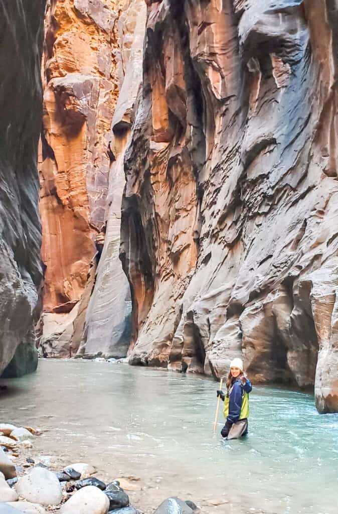 A woman standing in knee length water in the middle of a river in a slot canyon.