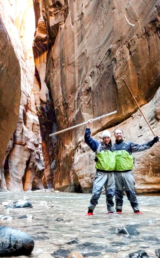 A man and woman stand in the middle of the Narrows, a large river leading throughout the large slot canyon.