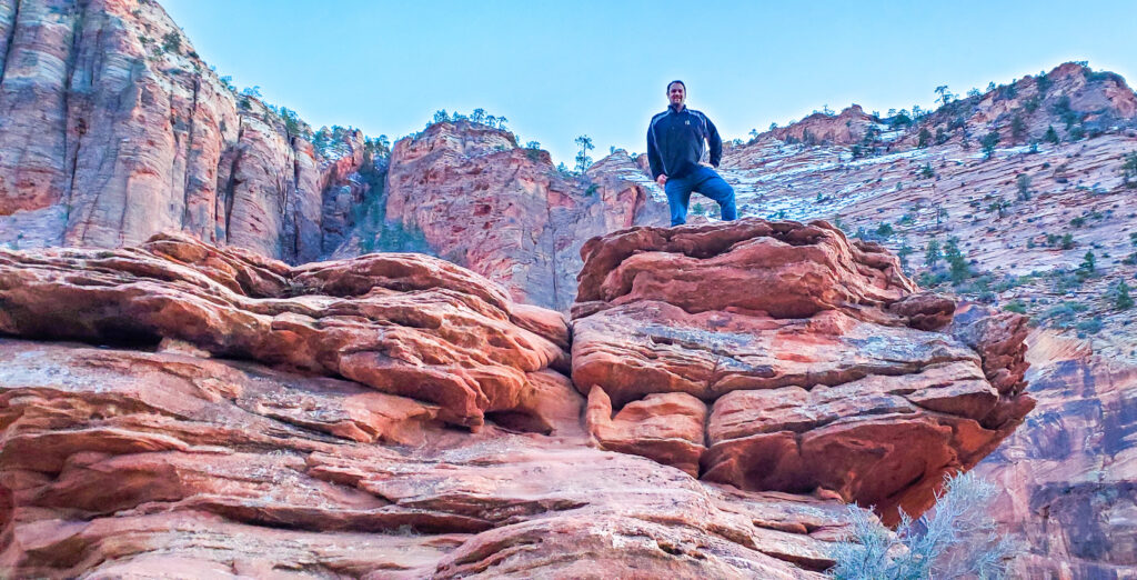 a man stands on top of a large dark orange rock formation with a large mountain in the background.