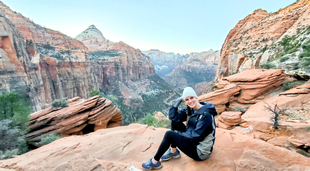 A woman sits on a large rock looking out at the large canyon with large colorful rock mountains in the background with trails running down into the canyon