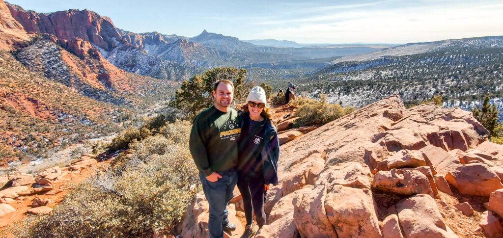 A man and woman stand on the rock overlook looking out at the large valley with lots of green pine trees and tall mountains on the left side.