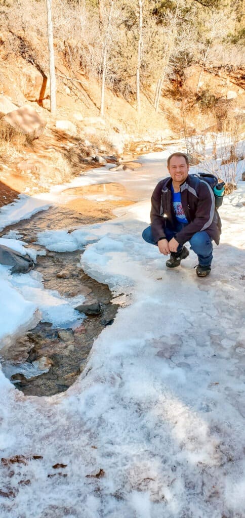 A man kneels on the ice in front of the base of a dirt mountain with an ice covered river in front.