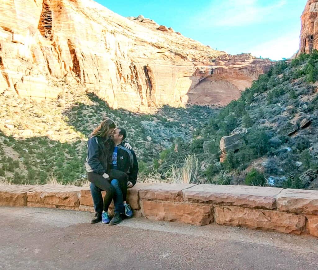 A woman sits on a man's lap at an overlook looking at the large rock walls with the rock formation curving around the valley.