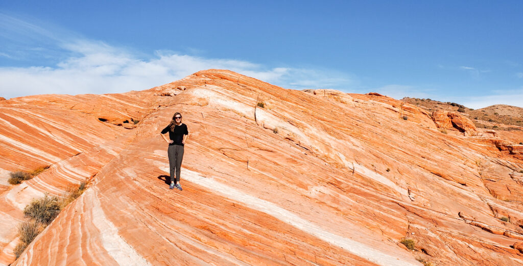 A woman stands on a large rock formation with large waves of different colored strata moving horizontally up the rock.