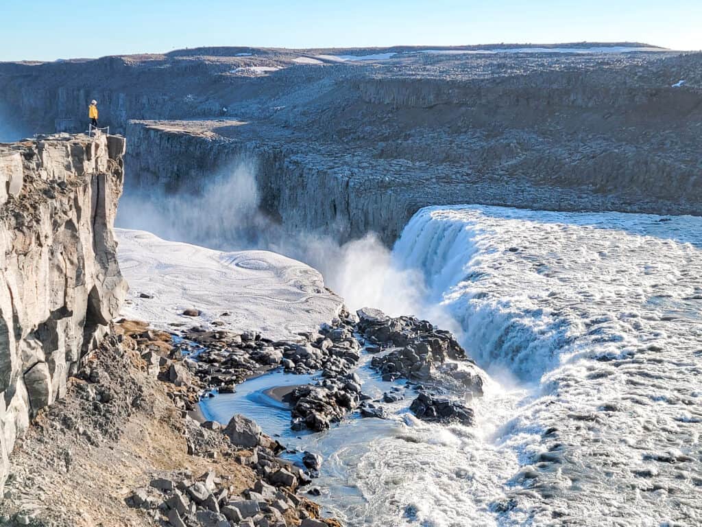 A woman stands on the edge of a cliff with white water crashes down a long way. Dettifoss is a huge waterfall and one of the best to see.