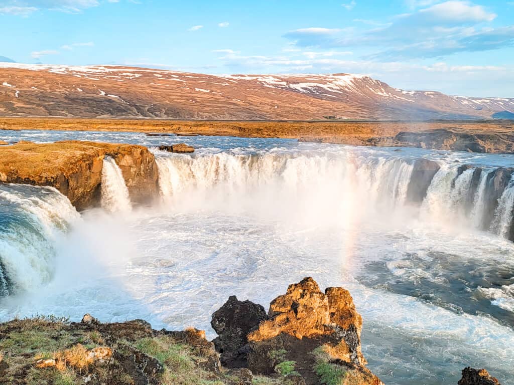 A very wide waterfall crashes down with a small mountain flat top in the background with a small rainbow.