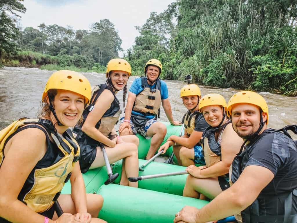 6 people sit on a white water raft, wearing life jackets and yellow helmets and smiling at the camera. You can see the rainforest behind them and the river in the background. Whitewater rafting is one of the funnest things to do in La Fortuna.