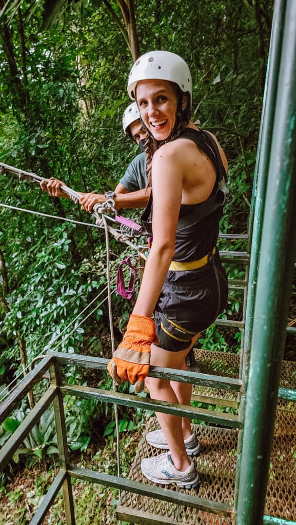 A woman in black, wearing a harness and a helmet, stands on a metal platform, hooked up to a rope. She is in the middle of the jungle.