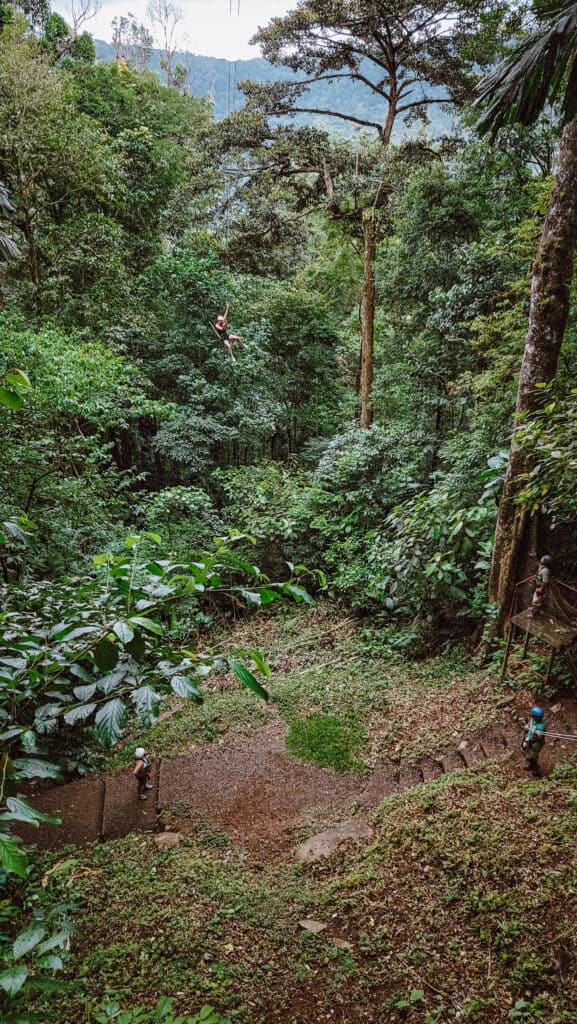 A wider shot of a rainforest clearing, with a woman swinging out in the distance on a rope swing. This is one of the funnest things to do in La Fortuna.