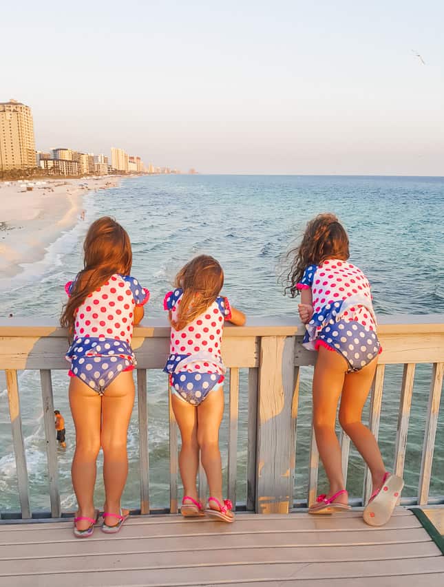 Three young girls in matching polka-dotted swimsuits look over the pier onto the water below. The waves of the ocean and part of the beach is in the background.