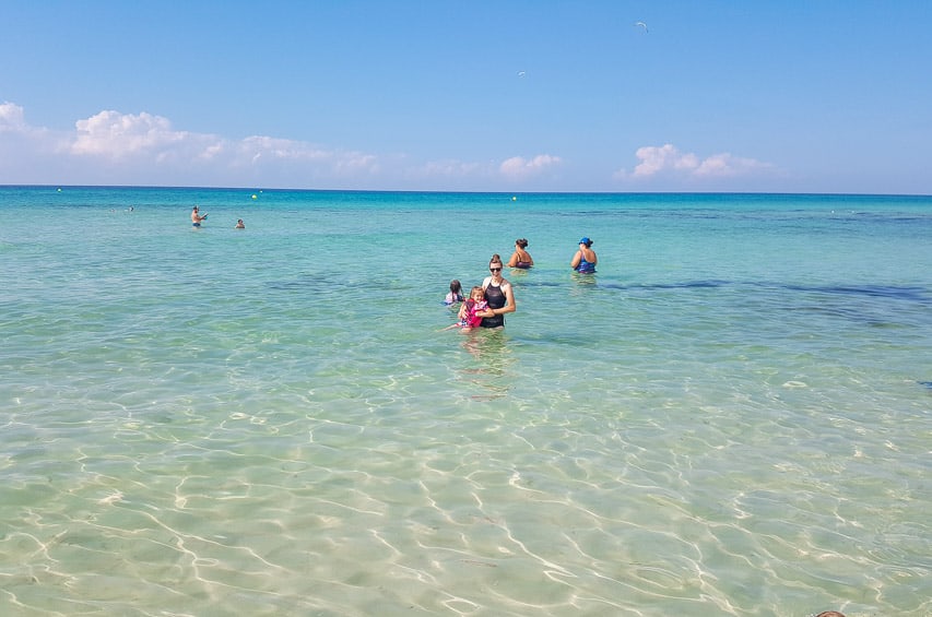 A woman in a black swim suit and a child in a pink life jacket stand up to their waist in the clear waters of the gulf coast.