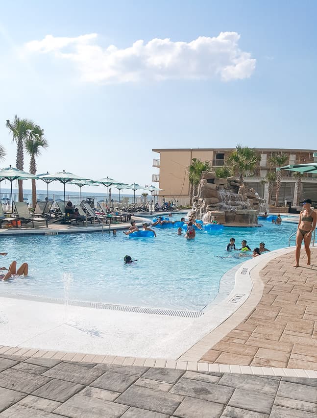 A swimming pool and deck are shown in the foreground - with 10-20 people playing in the water of the pool. In the background, some people are lounging on pool chairs and palm trees and the water of the ocean is shown behind them.
