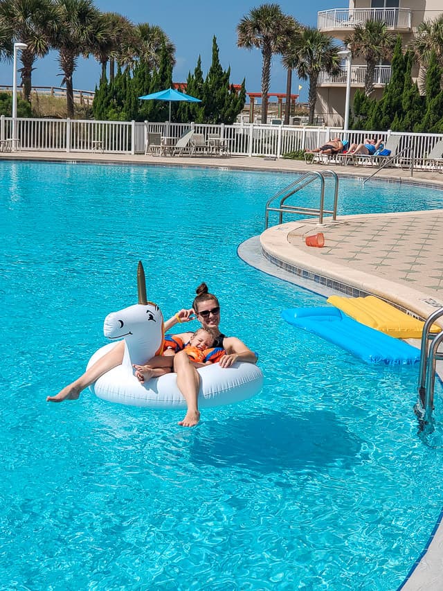 A woman and child are lying in a unicorn inner tube in the middle of a pool. The pool is very large and shaped like an 8, and some people lying on the pool loungers are visible in the background.