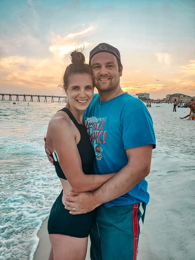 A couple is hugging and smiling at the camera as they stand on a beach at the edge of the water. The sky is full of colors from the sunset, and a pier extends into the water in the background.