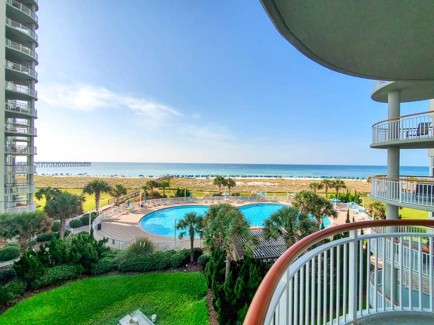 The view from a deck looking out over a pool area to the ocean. There are palm trees around the pool, grasses between the pool and the beach, and the deep blue ocean behind.