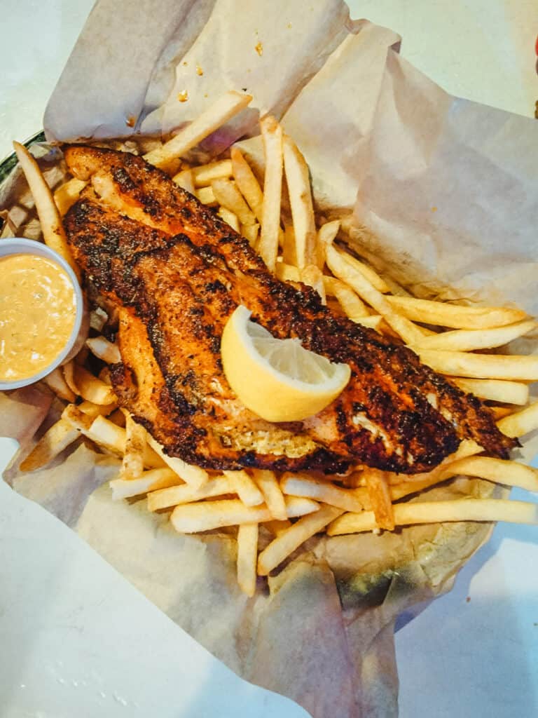 Blackened fish sits on top of a bed of french fries. There is dipping sauce in a little cup in the corner, and a wedge of lemon on top of the fish. The whole meal is served in a basket lined with brown paper.