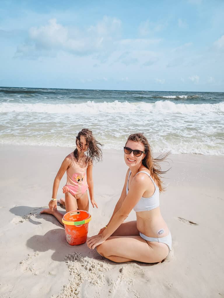 A woman in a white swimsuit and a girl in a pink swimsuit are on the beach, smiling at the camera, as they fill a pail with sand, with the waters of the ocean behind them.