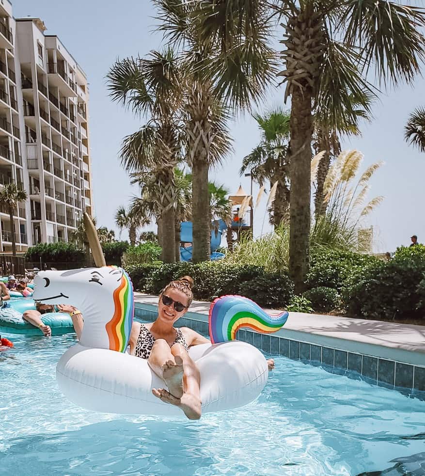 A woman in a cheetah print swimsuit and sunglasses is lying on a unicorn inner tube and smiling at the camera as she floats on a lazy river. Palm trees line the side of the pool.