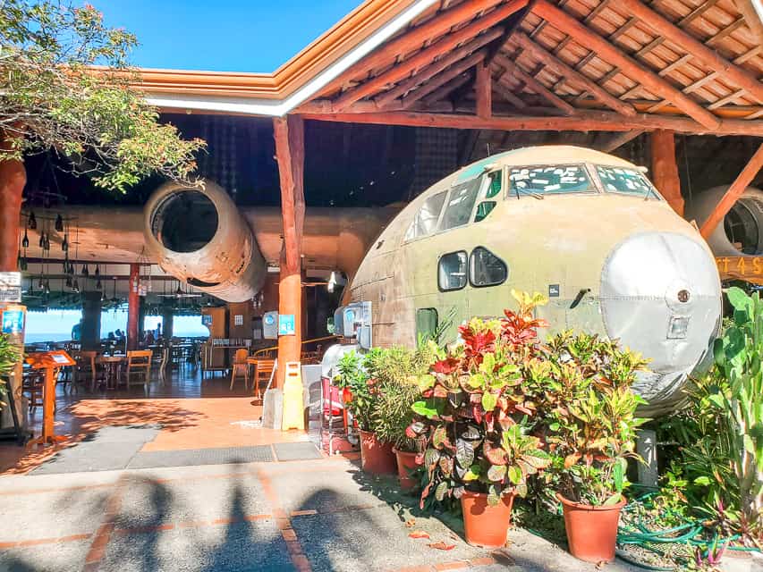 A restaurant is built around an old airplane. Potted plants decorate the sides of the airplane and you can see tables under the patio. The ocean is in the distance.