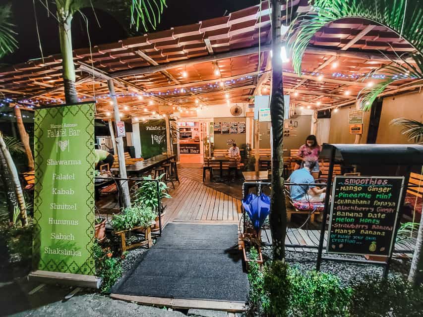 the patio of a restaurant is shown. There are low tables and reclining benches to sit on. It is night, and there are string lights on and palm fronds around.