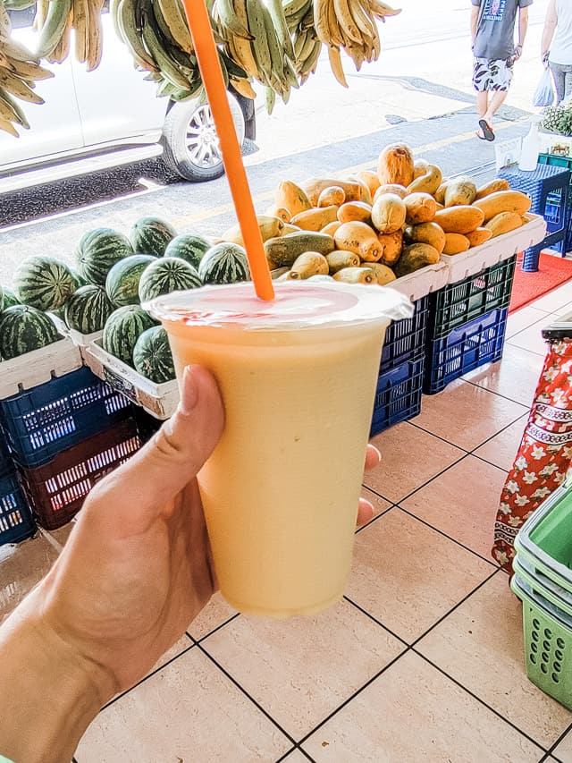 A hand is holding a mango smoothie in a plastic cup in a store. In the background are fruit displays from the store.
