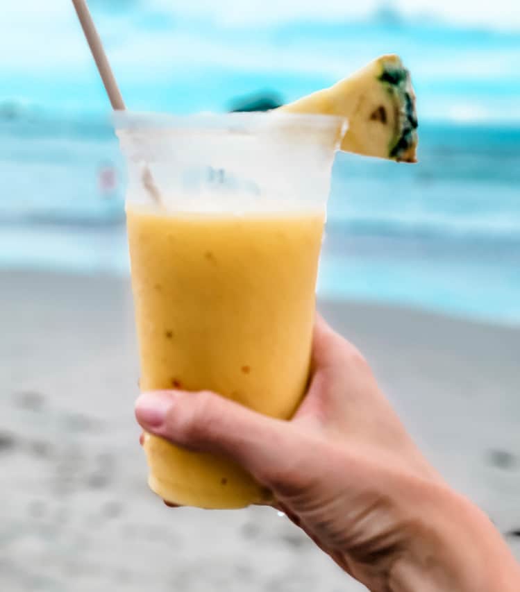 A yellow, mango smoothie is being held up on a beach, with sand and waves in the background. A straw and pineapple wedge are also on the cup.