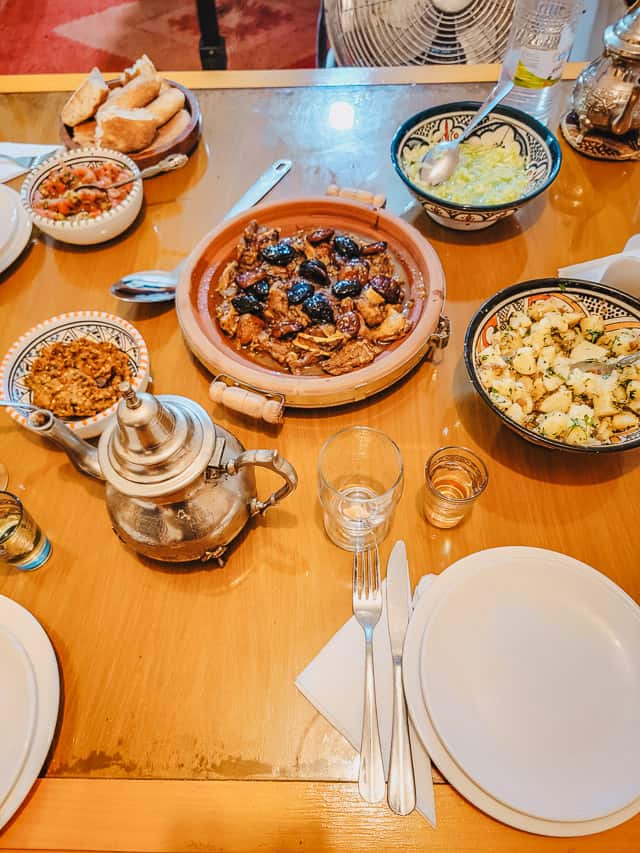 A table is set with white plates, silverware, and cups. A tagine with lamb and apricots is in the center, with a teapot, a bread bowl, and four smaller bowls of salads surrounds it.