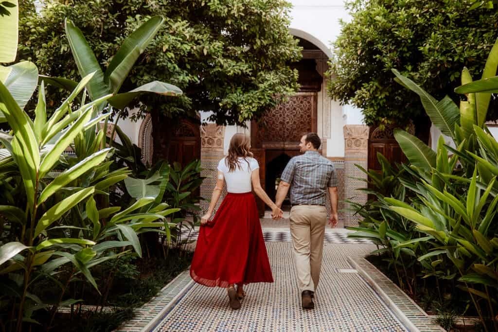 A man in a blue shirt and tan pants holds the hand of a woman in a white top and flowy, full-length red skirt. They walk along a colorful tiled path, with plants and trees lining the sides. Tiled archways are in the background.