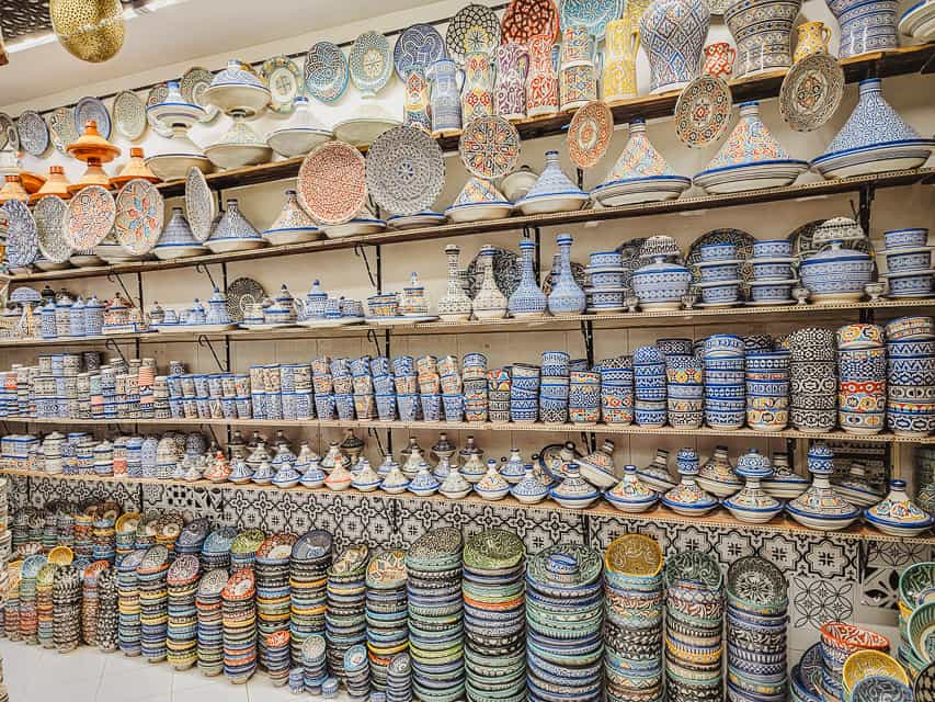 This view is the inside of a shop that is floor to ceiling filled with colorful ceramic bowls, plates, and tagines. Bowls are stacked high on the ground, and then there are four shelves filled with smaller bowls, small and large tagines, with large platters hung at the top of the walls.