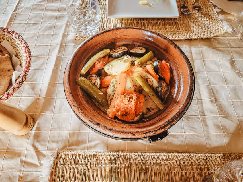 A chicken tagine is in the middle of a table with a cream tablecloth. Vegetables are stacked in the middle of the tagine. Wicker placemats, and a bread bowl are also partially visible.