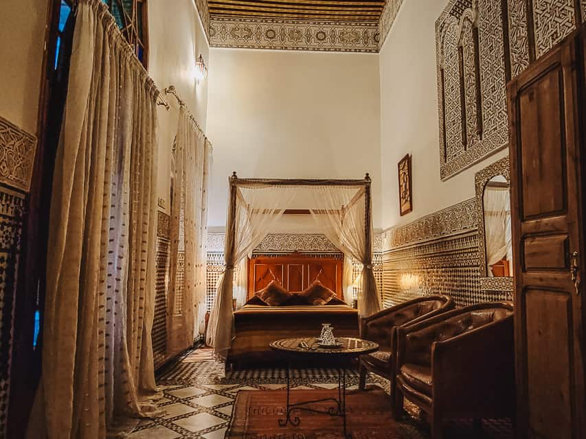 This is a room at riad Sara. There is a 4 poster bed with curtains drawn back. Colorful mosaics are on the walls and floor. The ceilings are extra high and have colorful wooden beams. Thick cream curtains line are in front of the doors and window, and two leather chairs in front of a small circular table are in the foreground.
