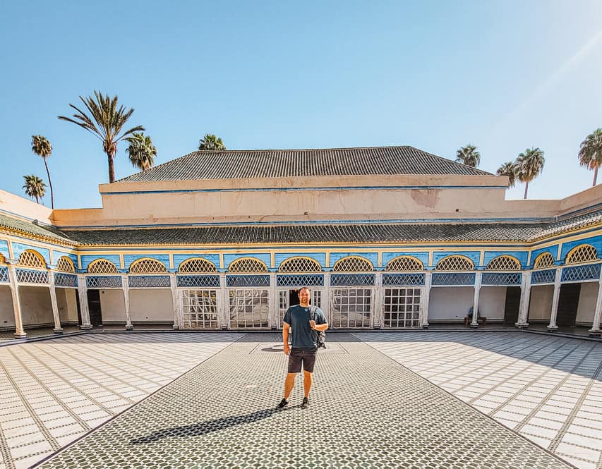 A man stands in the middle of the Grand Courtyard in the Bahia Palace, with white and black tiles on the floor, large, blue and white archways around the sides.