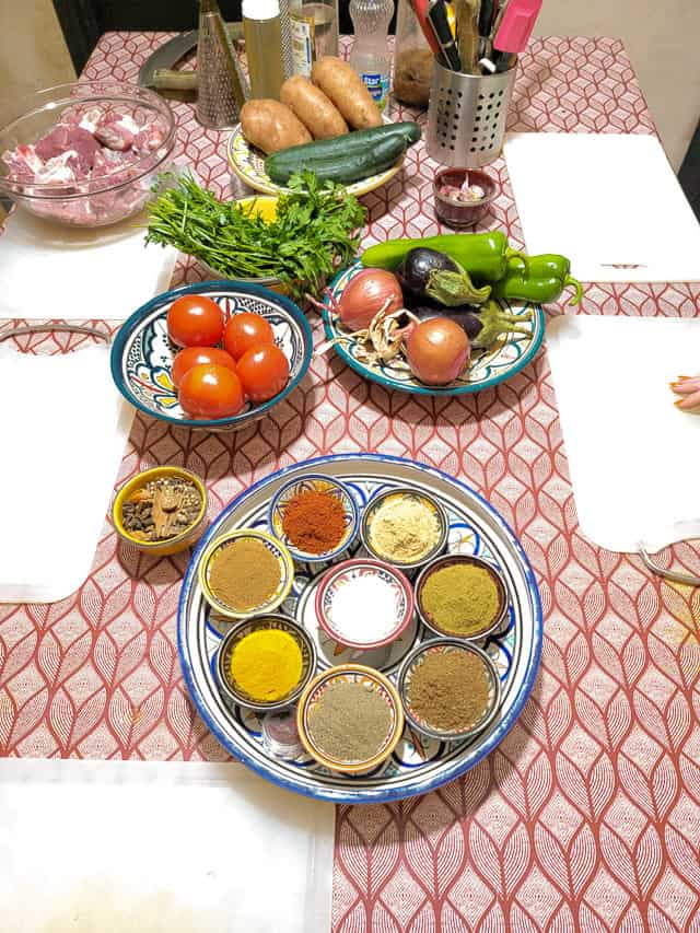 Plates of spices, vegeatbles, herbs, and meat are on a table that is set with cutttng boards