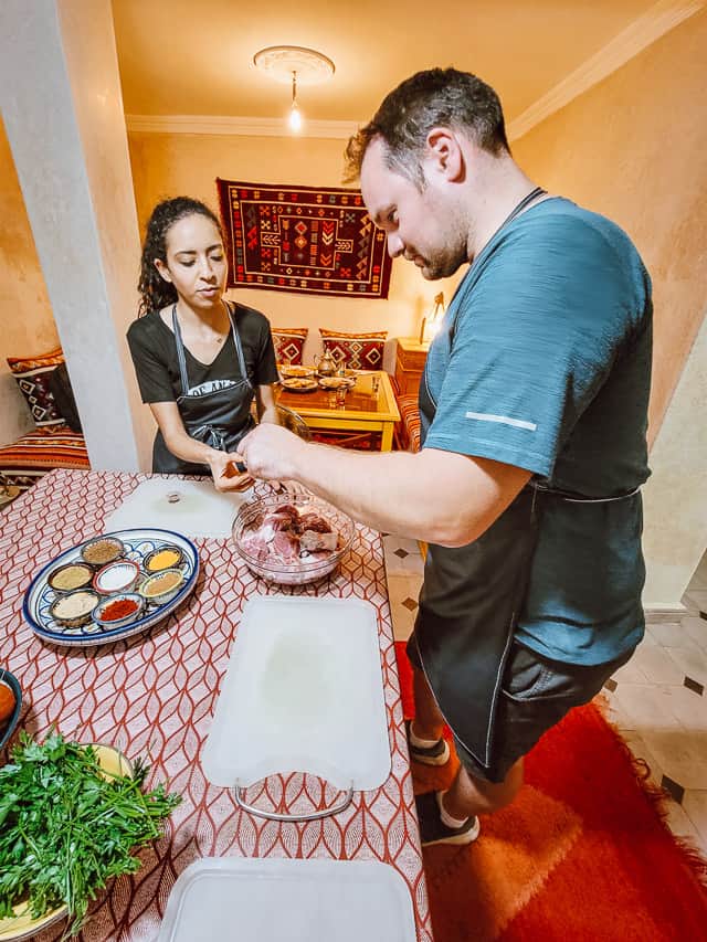 A Moroccan woman helps an American man measure spices to add to a bowl of lamb meat. They are both wearing black aprons. The table they are standing at has a red and white tablecloth, white cutting boards, and herbs and a plate of spices are visible. A colorfully red seating area is in the background.