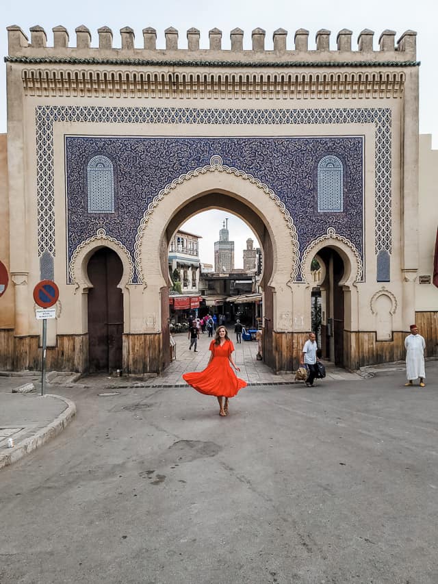 A woman in a red dress stands in the center of the Blue Gate arch. A few people are coming and going to the sides of the gate. The gate is covered in blue pottery.
