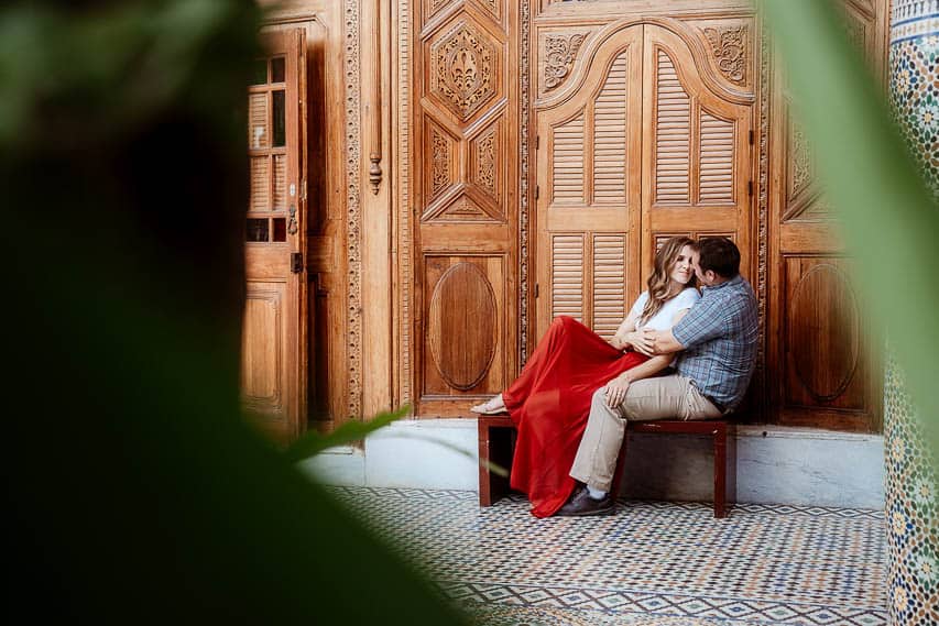 A man and a woman are sitting on a bench and smiling at each other in the musee dar el bacha. The view is partially bewteen leaves of plants in the musuem.