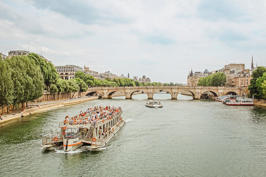 A sightseeing boat cruises down the Seine River, which is lined by walkways and trees. In the background is a bridge.