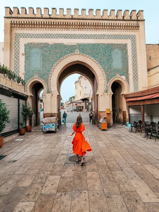 A woman in a red dress walks towards the green side of the Blue Gate. Tables from a restaurant are visible on the sides of the photo.