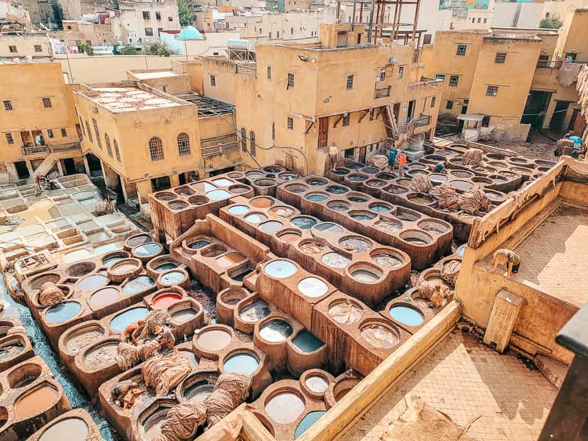 An overhead view down into the tanneries of Fes - dozens of large, round vats are filled with liqiuds of various colors, with stacks of leather skins on piled near some of the vats.