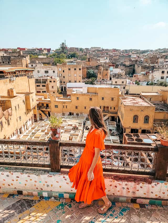 A woman in a red dress stands on a terrace and looks down at the basins filled with colored liquids in the leather tannery factory. In the background are the buildings and rooftops of Fes.