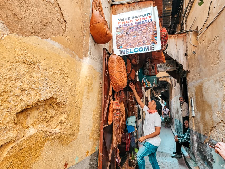 A narrow street with deep beige walls. The entrance to a shop is prominent in the forefront. A sign says "free visit", a worker is standing outside, and leather poufs and purses and displayed around the door.
