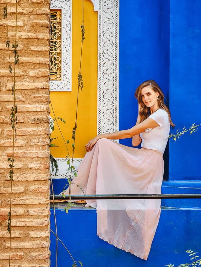 A woman in a white shirt and pink skirt sits on the ledge of a building painted deep blue and vibrant yellow in the Jardin Majorelle, a uniquely designed building in Marrakech.